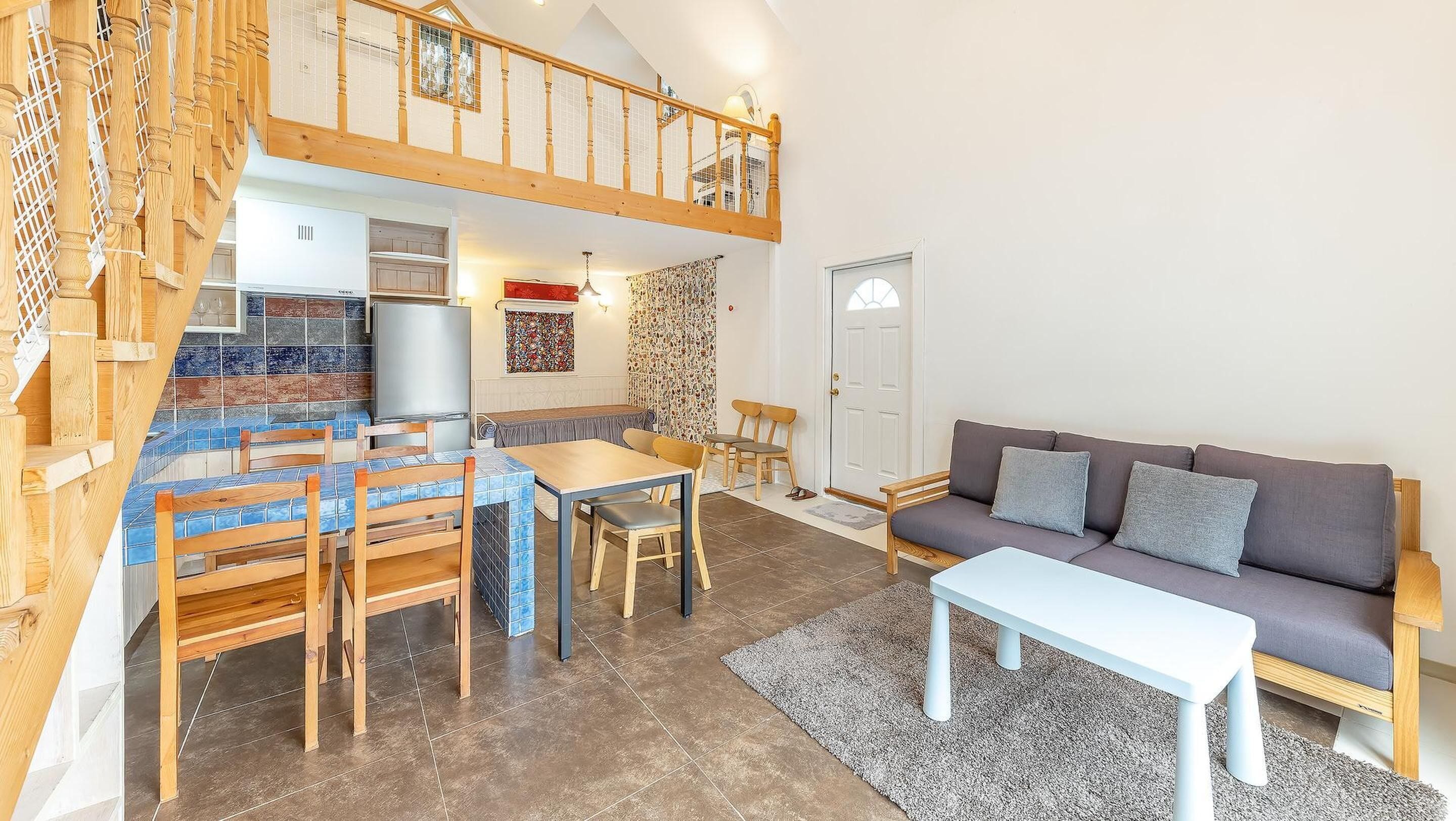 A spacious living area with a high ceiling, a dining table with chairs, a sofa, and a staircase with a wooden railing.