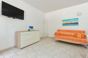Ett rum med en platt-TV som är monterad på väggen, ett vitt skåp, en orange soffa, en tavla som föreställer en solnedgång och en klocka på väggen.