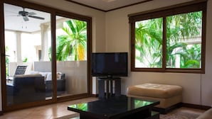Sala de estar com televisão de tela plana, mesa de centro de vidro, sofá e janela com vista para palmeiras.