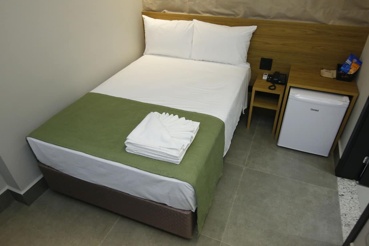 A hotel room with a bed, a nightstand, a small refrigerator, and a green bedspread.
