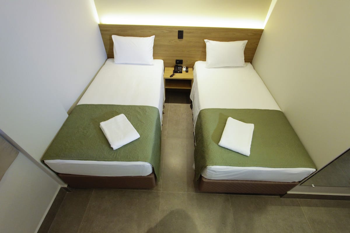 A hotel room with two single beds, each with a green bedspread and white pillows. There is a wooden headboard, a nightstand with a phone, and a small table between the beds.