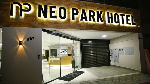 The exterior of the Neo Park Hotel with a prominent illuminated sign, a reception area visible through glass doors, and a numbered entrance door. - NEO PARK HOTEL (Maringá)