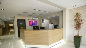 A modern reception desk with a wooden finish, a flat-screen TV mounted on the wall, and a large potted plant to the right. - NEO PARK HOTEL (Maringá)
