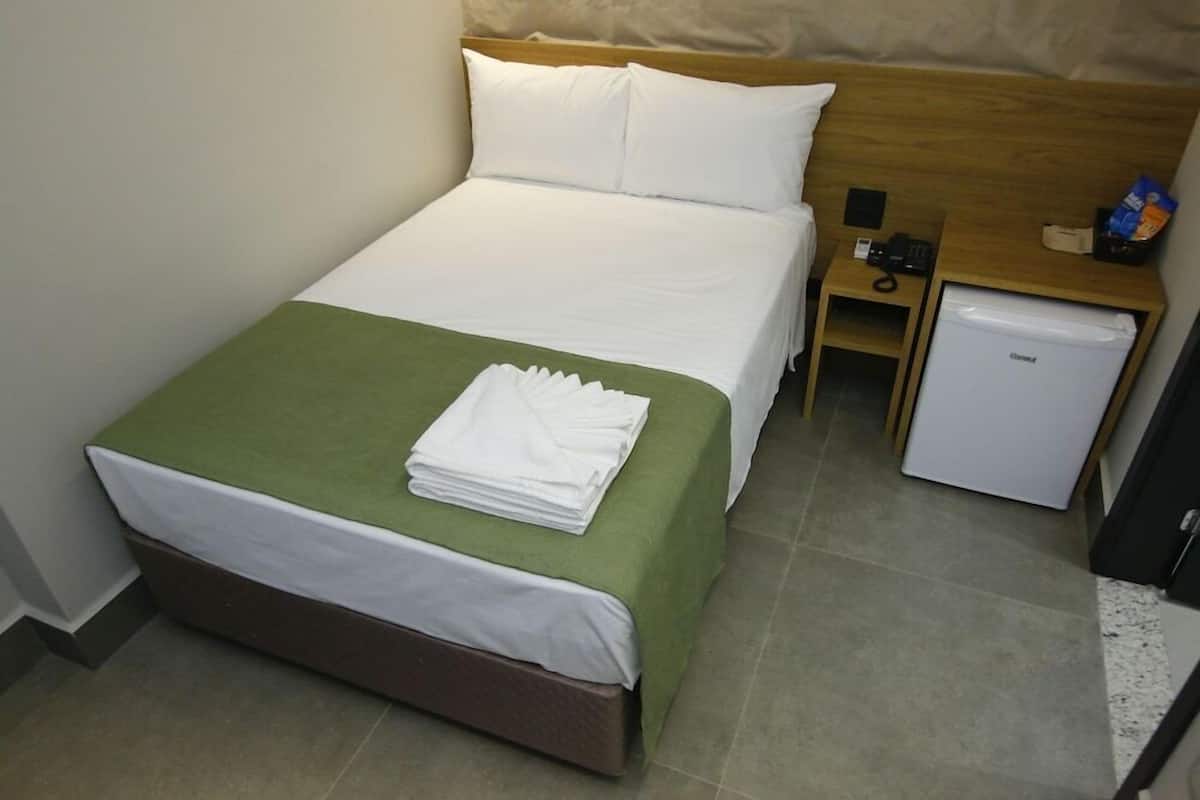 A hotel room with a bed, green bedspread, white pillows, a small bedside table with a phone, and a mini fridge.