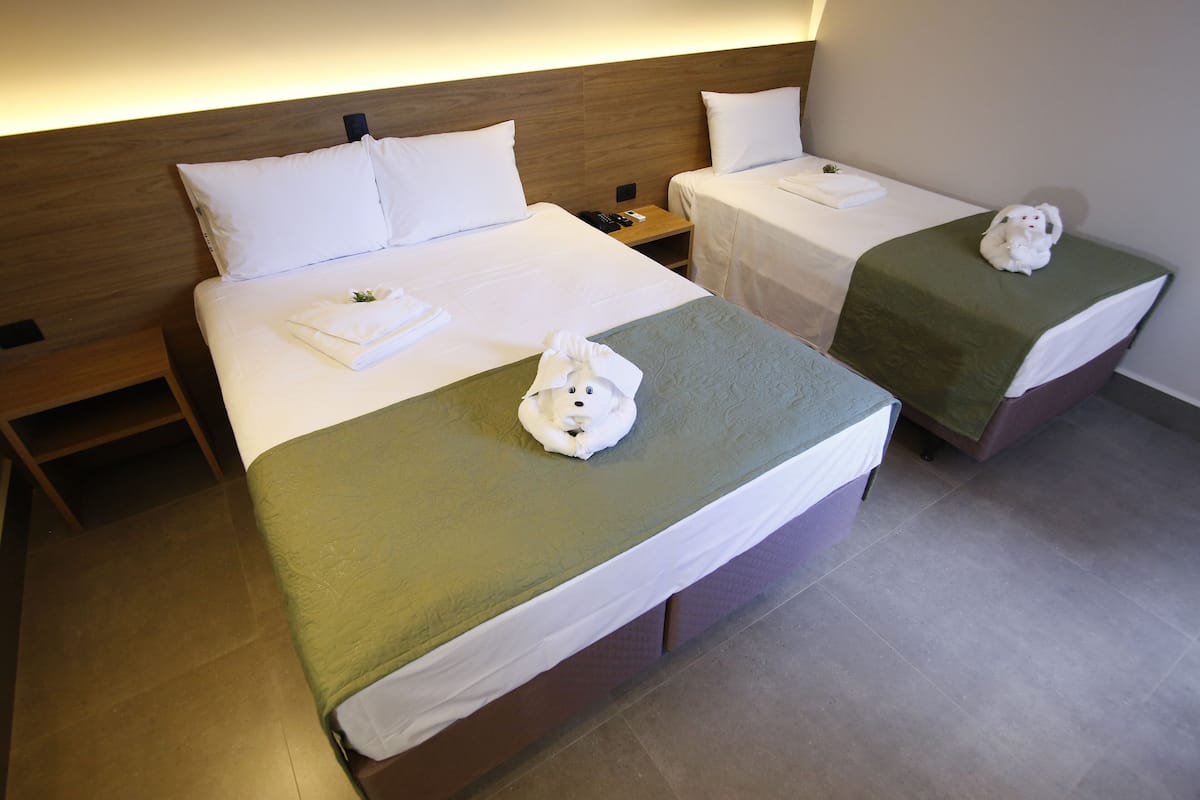 A hotel room with two beds, each with a green bedspread and white pillows. There are towels on the beds and a small nightstand with a phone and remote control.