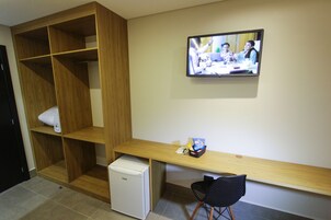 A hotel room with a wooden wardrobe, a small white refrigerator, a wooden desk with a chair, and a television mounted on the wall.