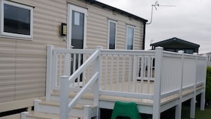 Una casa móvil con terraza blanca, una silla de plástico verde y una escalera que lleva a la entrada.
