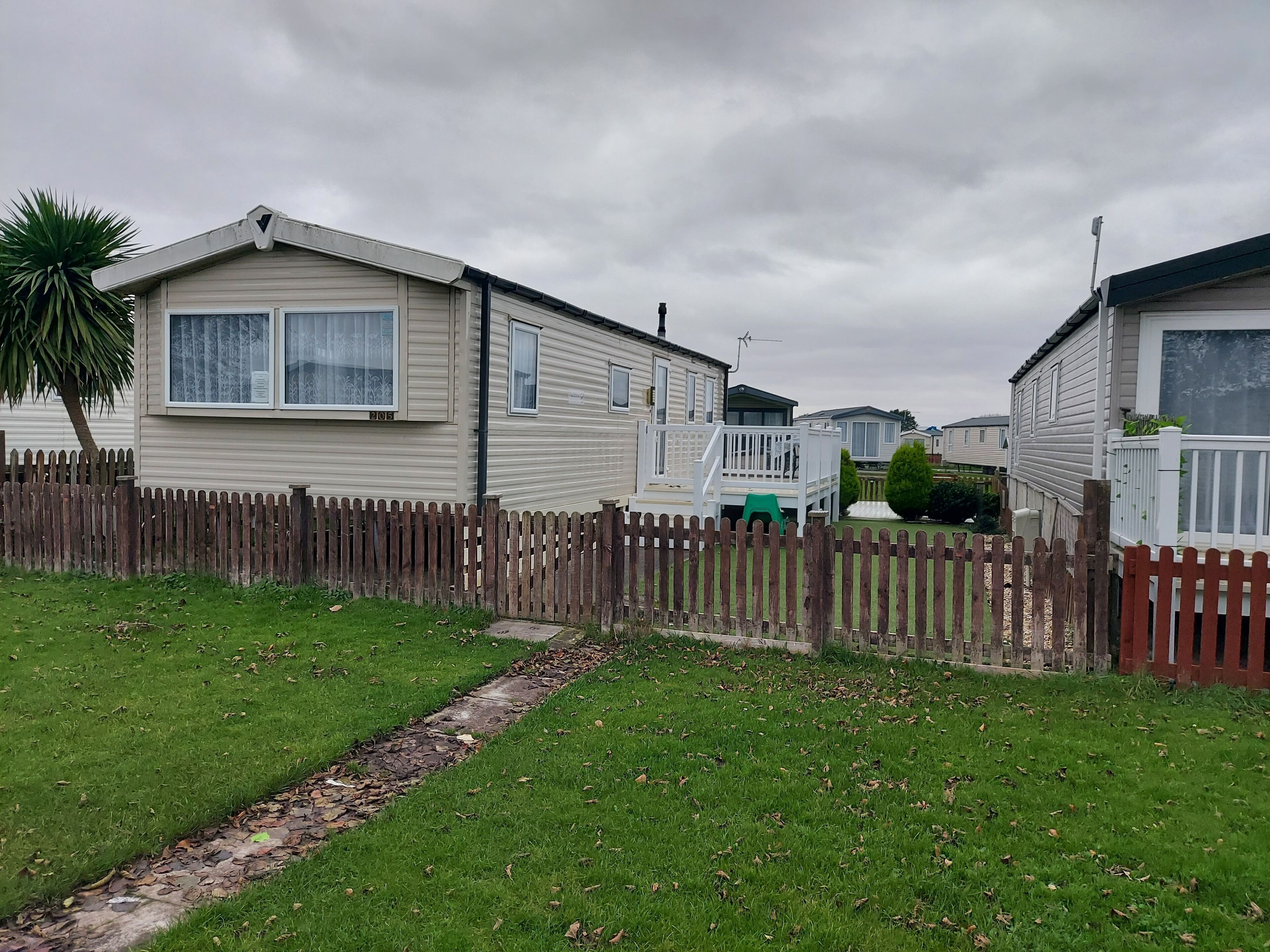 A row of mobile homes with a wooden fence in front, a grassy area, and a cloudy sky.