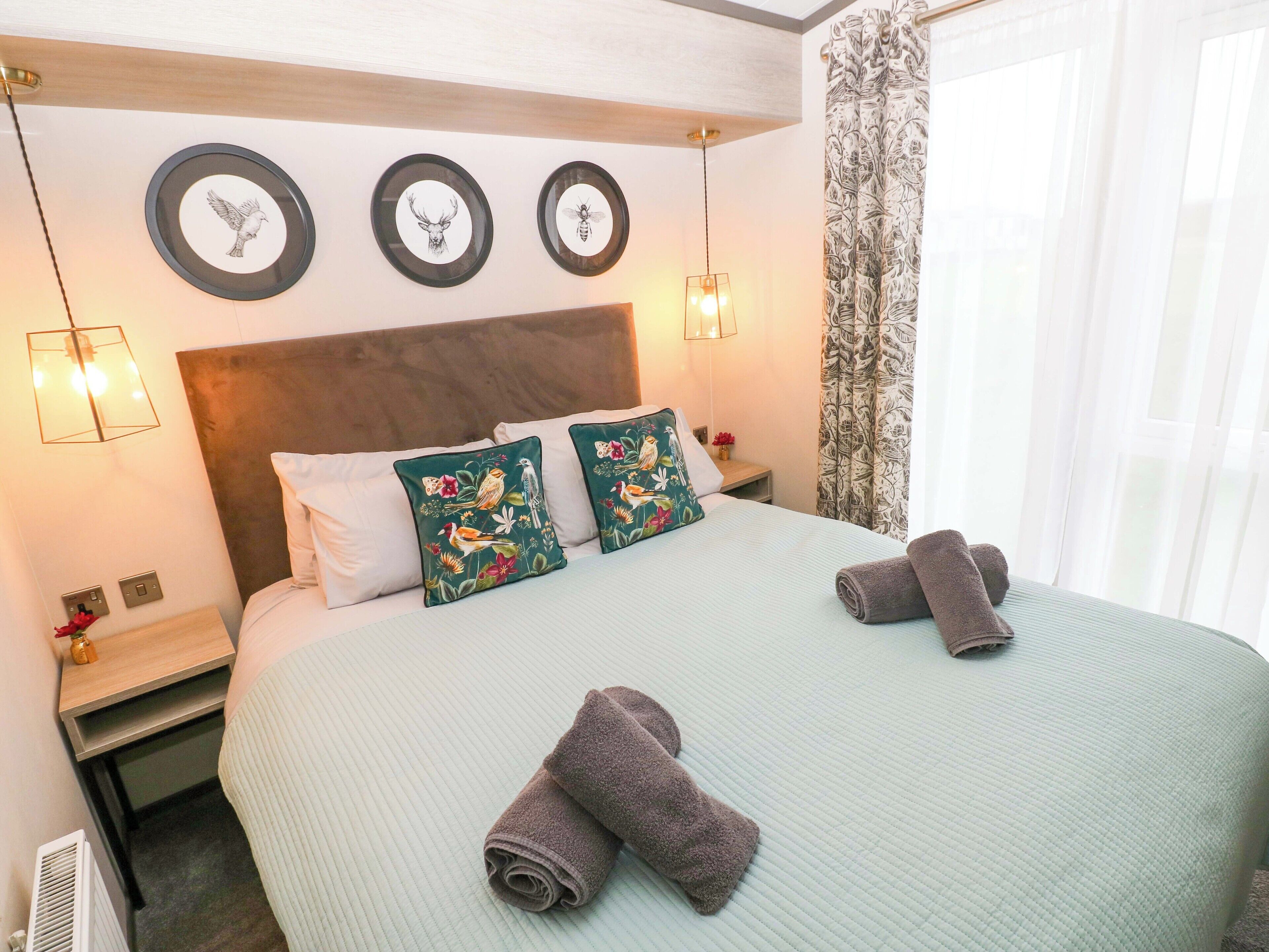 A hotel room with a bed, two pillows with floral designs, a headboard, bedside lamps, and wall art depicting birds.