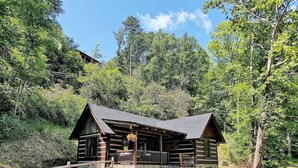 A log cabin with a porch and stone foundation, surrounded by dense forest and a clear sky. - CastAway Waterfront Rustic Cabin Very Private With Outdoor Cabana (Waynesville)