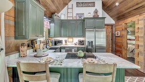 A kitchen with green cabinets, a central island with chairs, a stainless steel refrigerator, and a 'Welcome' sign on the wall. - CastAway Waterfront Rustic Cabin Very Private With Outdoor Cabana (Waynesville)