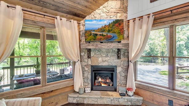 A cozy room with a stone fireplace, large windows with curtains, a comfortable sofa, and a scenic painting above the fireplace. - CastAway Waterfront Rustic Cabin Very Private With Outdoor Cabana (Waynesville)