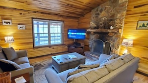 A cozy cabin interior with wooden walls, a stone fireplace, a television, and a large window. - Secluded, Great View! Hot Tub, Wood Fireplace, Screened in Porch, Arcade Games. (Rogers)