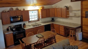 A compact kitchen with wooden cabinets, a microwave, stove, and a dining area with a table and chairs. - Secluded, Great View! Hot Tub, Wood Fireplace, Screened in Porch, Arcade Games. (Rogers)