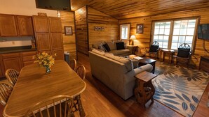 A cozy cabin interior with a wooden dining table, sofa, and chairs, a small kitchen area, and a large window providing natural light. - Secluded, Great View! Hot Tub, Wood Fireplace, Screened in Porch, Arcade Games. (Rogers)