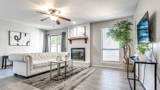 A spacious living room with a white sofa, a fireplace, a ceiling fan, and a large window with blinds.