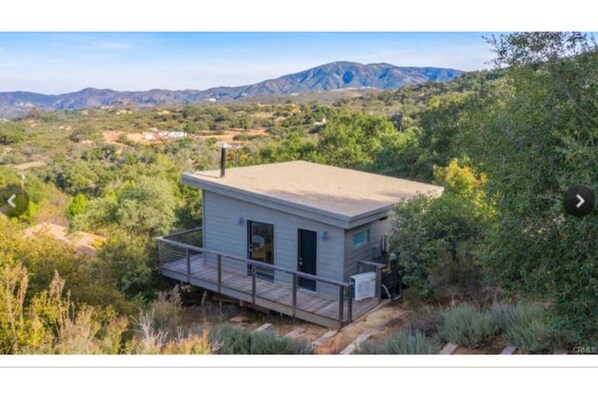 A small, single-story cabin with a deck, surrounded by dense vegetation and mountains in the background.