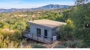 A small, single-story cabin with a deck, surrounded by dense vegetation and mountains in the background.