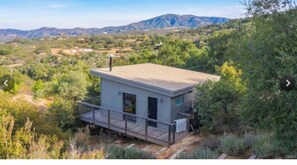 A small, single-story cabin with a deck, surrounded by dense vegetation and mountains in the background.