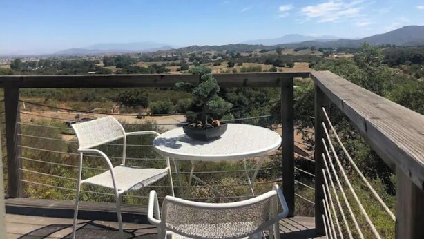 Varanda com mesa e duas cadeiras, com vista para uma paisagem deslumbrante com montanhas e campos.