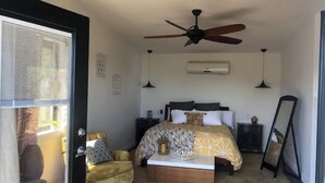 Quarto com cama, ventilador de teto, ar condicionado, mesa de cabeceira, cadeira e espelho.