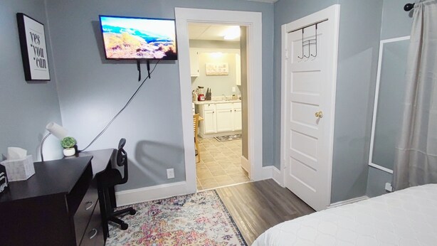 Un dormitorio con una cama, un escritorio con silla, un televisor montado en la pared y una puerta que da a un baño.