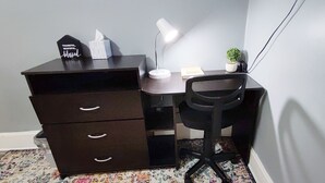 Un pequeño escritorio oscuro con una silla, una lámpara, una caja de pañuelos y una planta pequeña en maceta.