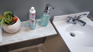 Un baño con lavamanos blanco, grifo cromado, toalla blanca, jarra blanca con una planta y dos botellas de jabón líquido.