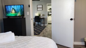 Habitación de hotel con una cama, un televisor que muestra una escena de bosque, un escritorio con una silla y una puerta que da a otra habitación.