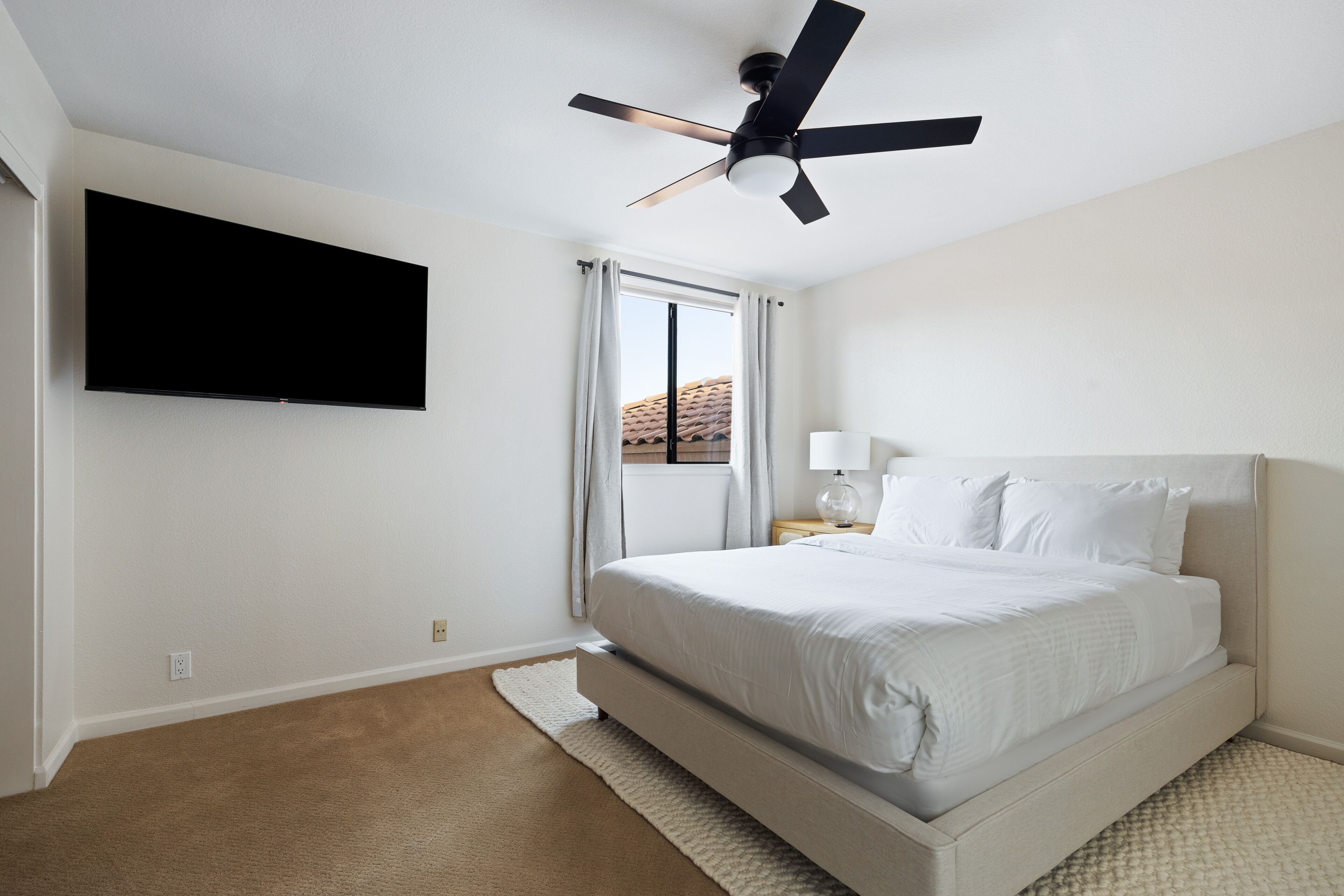 Une chambre à coucher avec un lit, un ventilateur de plafond, un téléviseur fixé au mur et une fenêtre avec des rideaux.