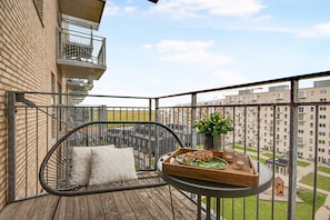 Un balcon avec un fauteuil en osier, une petite table sur laquelle est posé un plateau de nourriture, et une vue sur les bâtiments environnants.