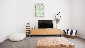 Olohuoneessa on puinen TV-taso, litteänäyttöinen televisio, punottu rahi, valkoinen koristetyyny, raidallinen maljakko, koristeellinen kasvi ja seinällä oleva taideteos kehyksissä.