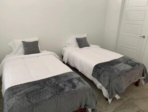 Two single beds with white mattresses and grey quilted covers, set against a plain white wall.