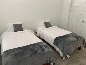Two single beds with white mattresses and grey quilted covers, set against a plain white wall.