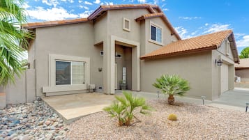 A two-story house with a tiled roof, a front porch with a bench, and a small garden with palm trees and a cactus.