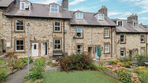 A row of stone houses with multiple windows and doors, a small garden with various plants and flowers, and a clear blue sky. - Longstone View (Bakewell)