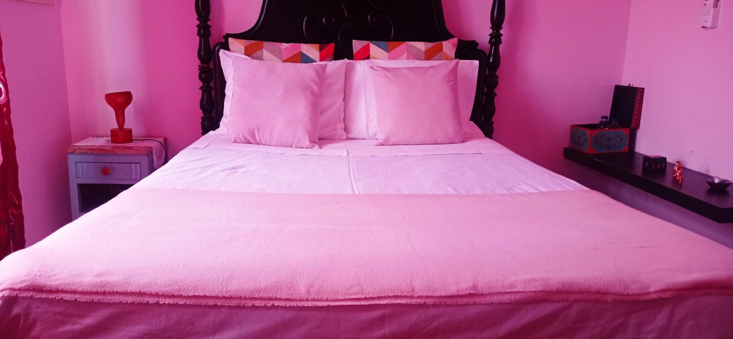 Una cama con cabecera negra, ropa de cama rosa y un cojín de colores.