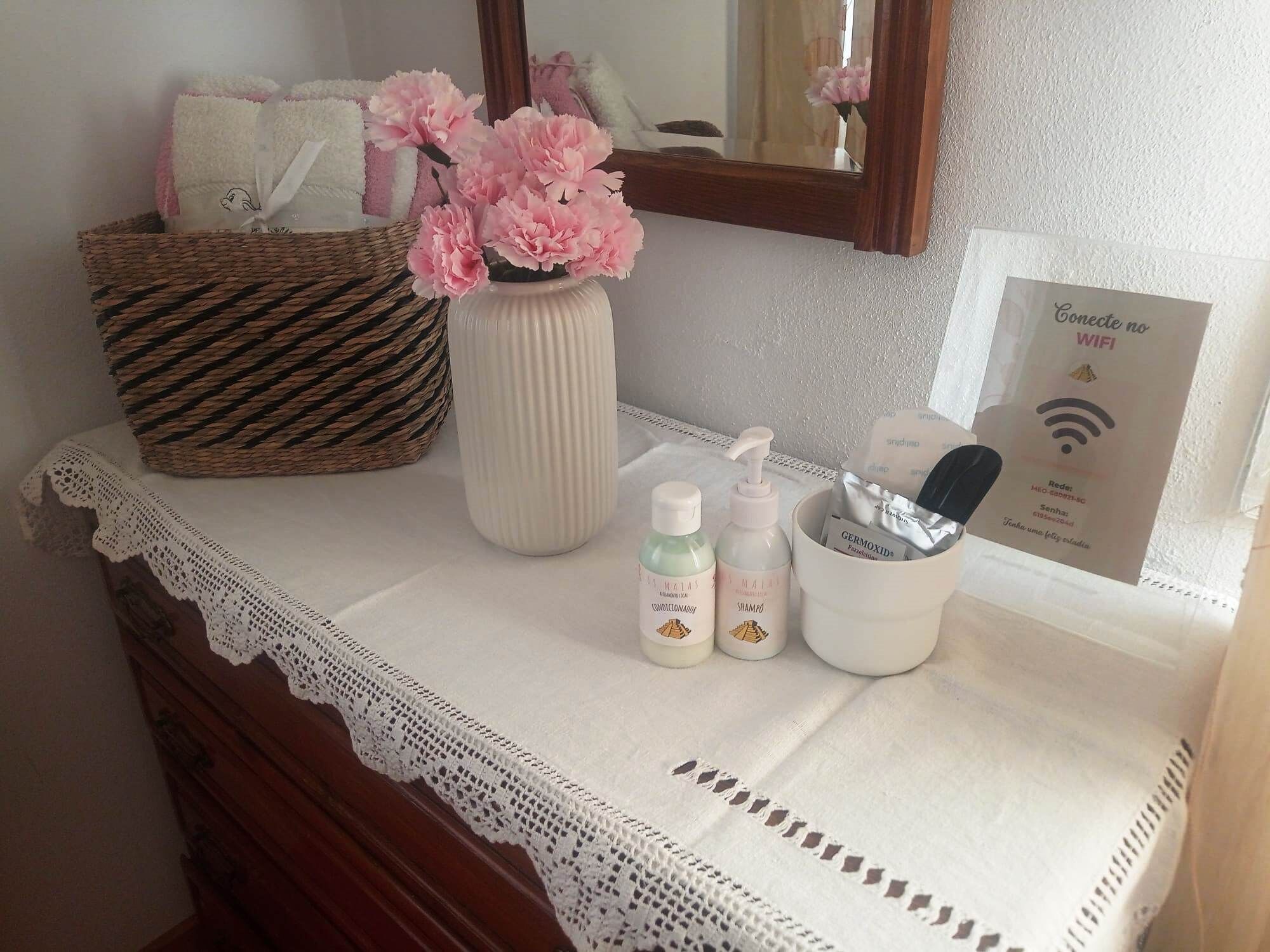 A bathroom vanity with a white lace doily, a woven basket, a vase with pink flowers, and toiletries.