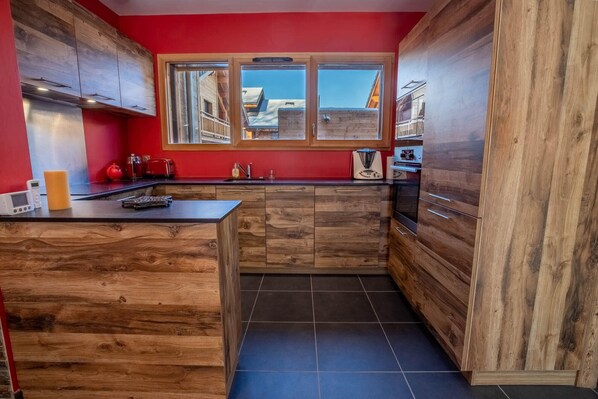 Una cocina con armarios de madera, una isla central, una pared roja y un ventanal con vista a edificios.