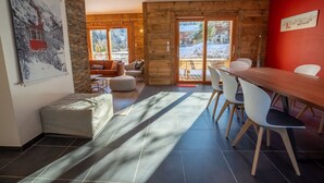 A cozy room with a wooden interior, a dining area with white chairs, and a large window offering a view of a snowy landscape. - Chalet de l'Olympe A2 (Les Allues)