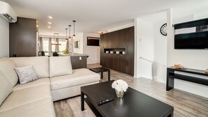 A modern living room with a sofa, a coffee table with a vase of flowers, a kitchen area with a built-in oven, and a TV mounted on the wall. - Peaceful House 10 minutes to ottawa and Excellent location (Gatineau)