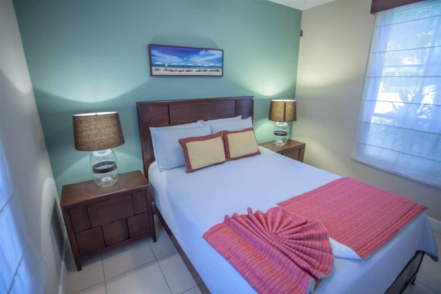 Habitación de hotel con cama, dos mesitas de noche, dos lámparas, una ventana con persianas y un cuadro en la pared.