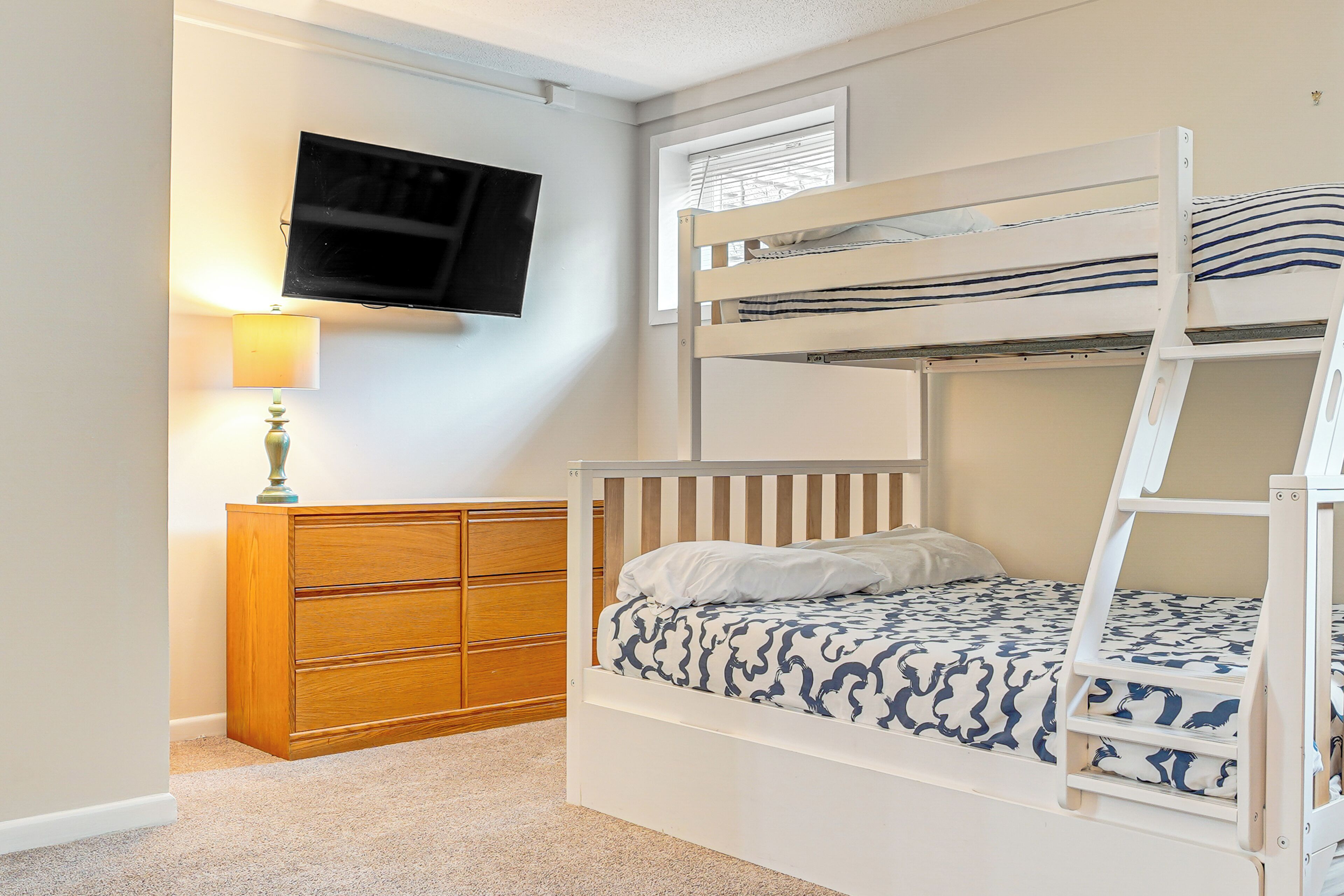 A bunk bed with a TV mounted on the wall, a wooden dresser, and a window with blinds.