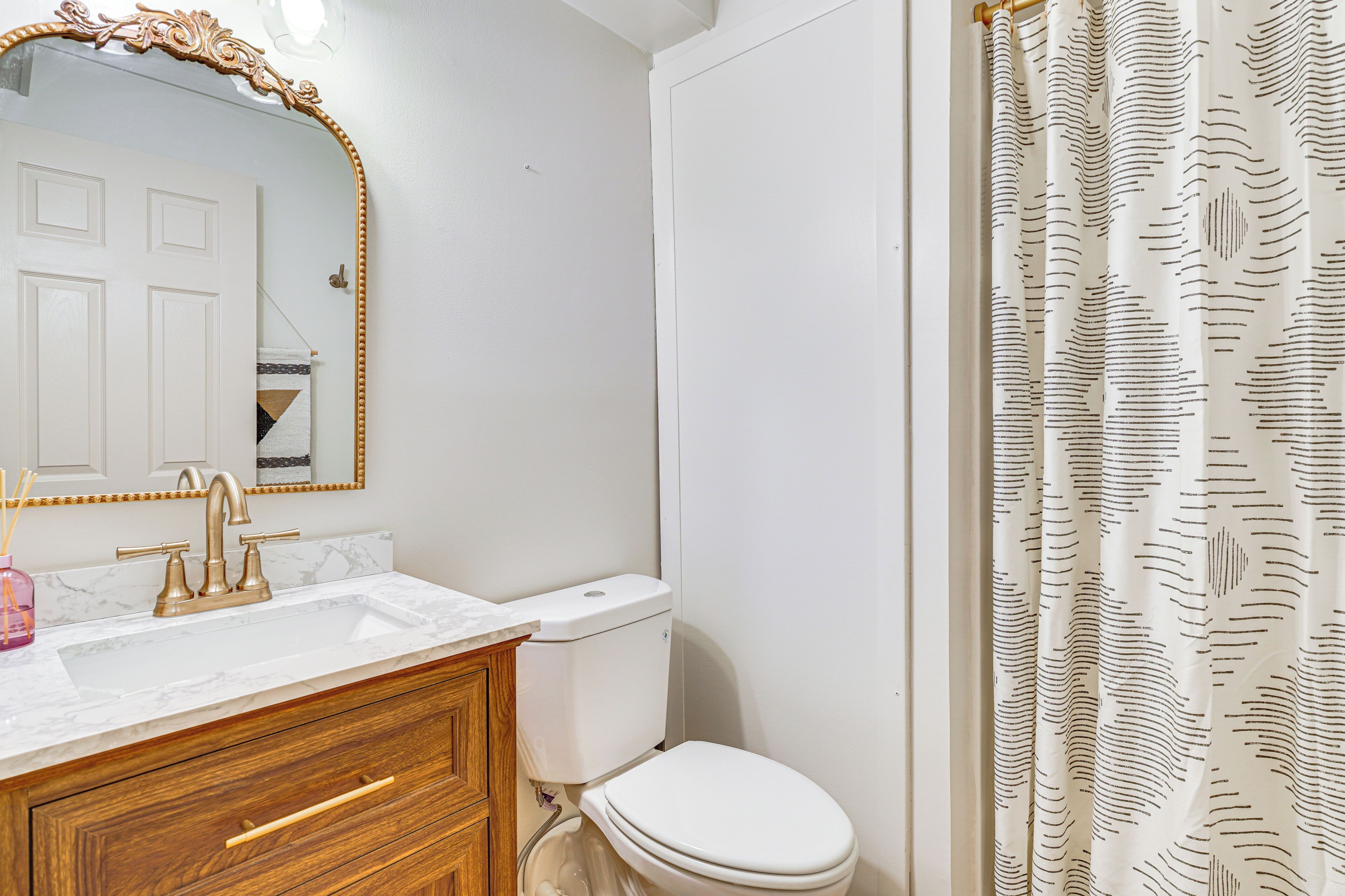 A bathroom with a white sink, gold faucets, a wooden vanity, a white toilet, and a shower curtain with a wavy pattern.