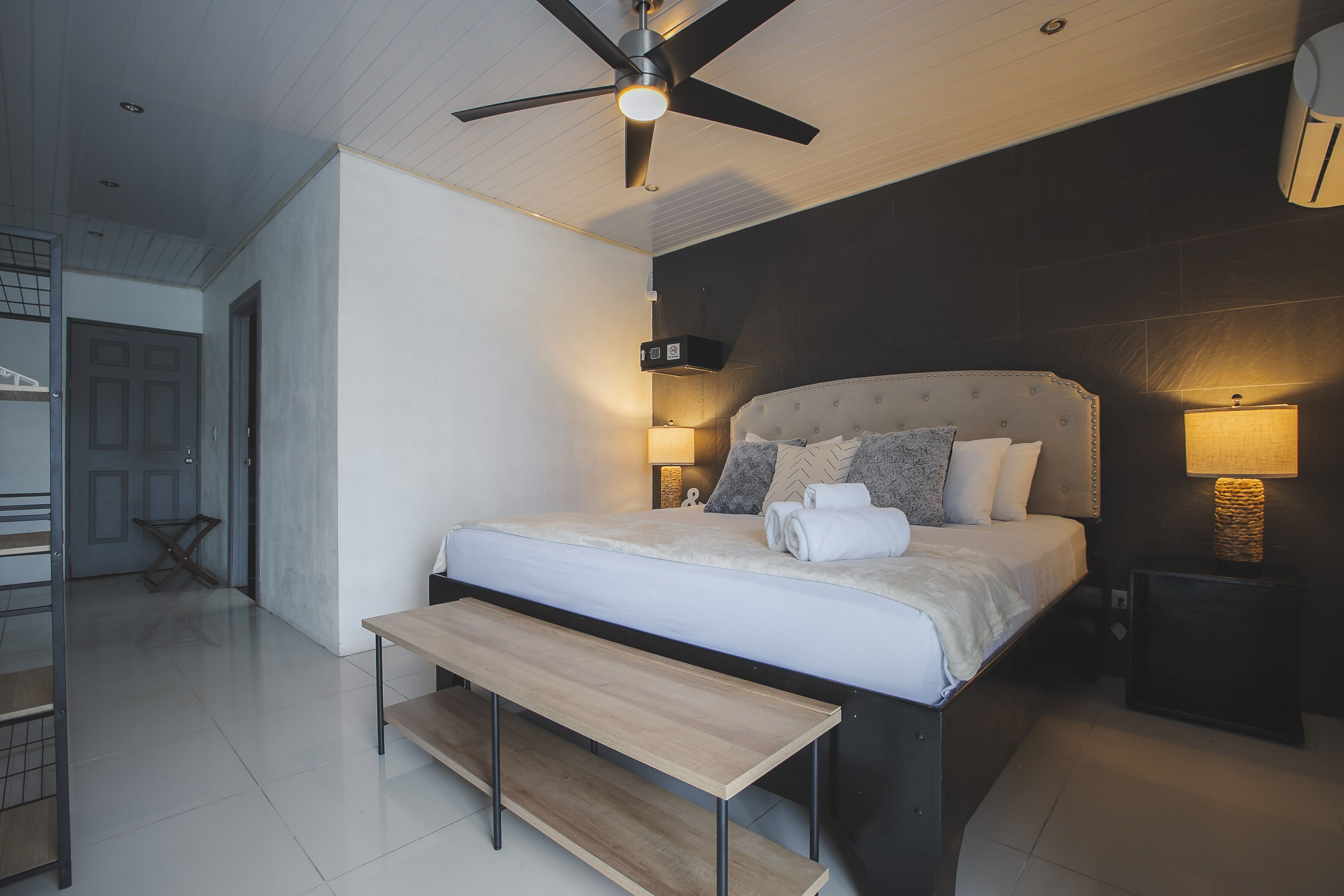 Un dormitorio moderno con una cama grande, un ventilador de techo, lámparas de noche y un banco.
