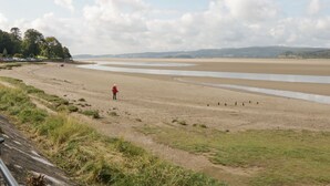 A beach with a person in a red jacket walking along the shore. - Rolo's Retreat (Grange-over-Sands)