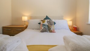 A neatly made bed with white linens, a tropical leaf-patterned pillow, and a bedside table with a lamp and a tissue box. - Rolo's Retreat (Grange-over-Sands)