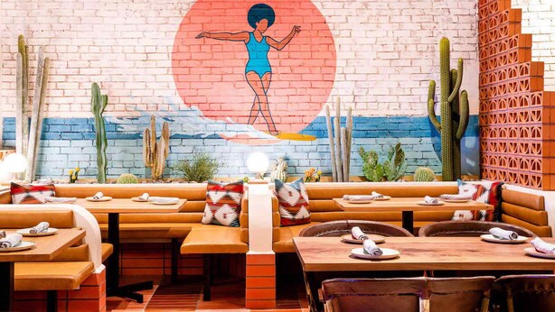 Un restaurante con mesas y bancos de madera, un mural en la pared y decoración de cactus.
