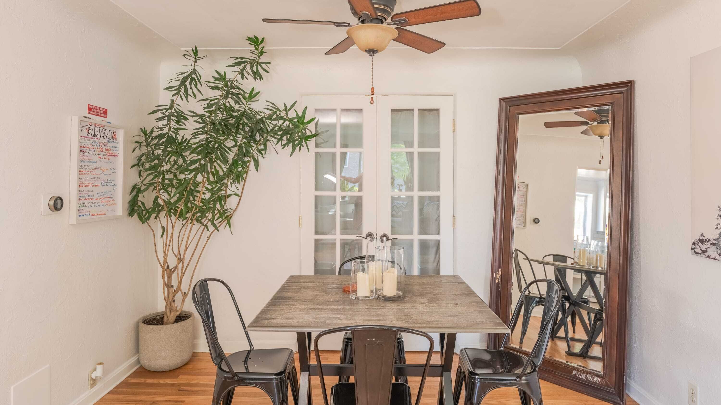 Un coin repas avec une table en bois, quatre chaises, un ventilateur de plafond, un grand miroir et une plante en pot.