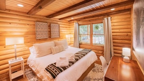 A log cabin bedroom with a bed, two bedside tables, a desk, and a window with curtains.